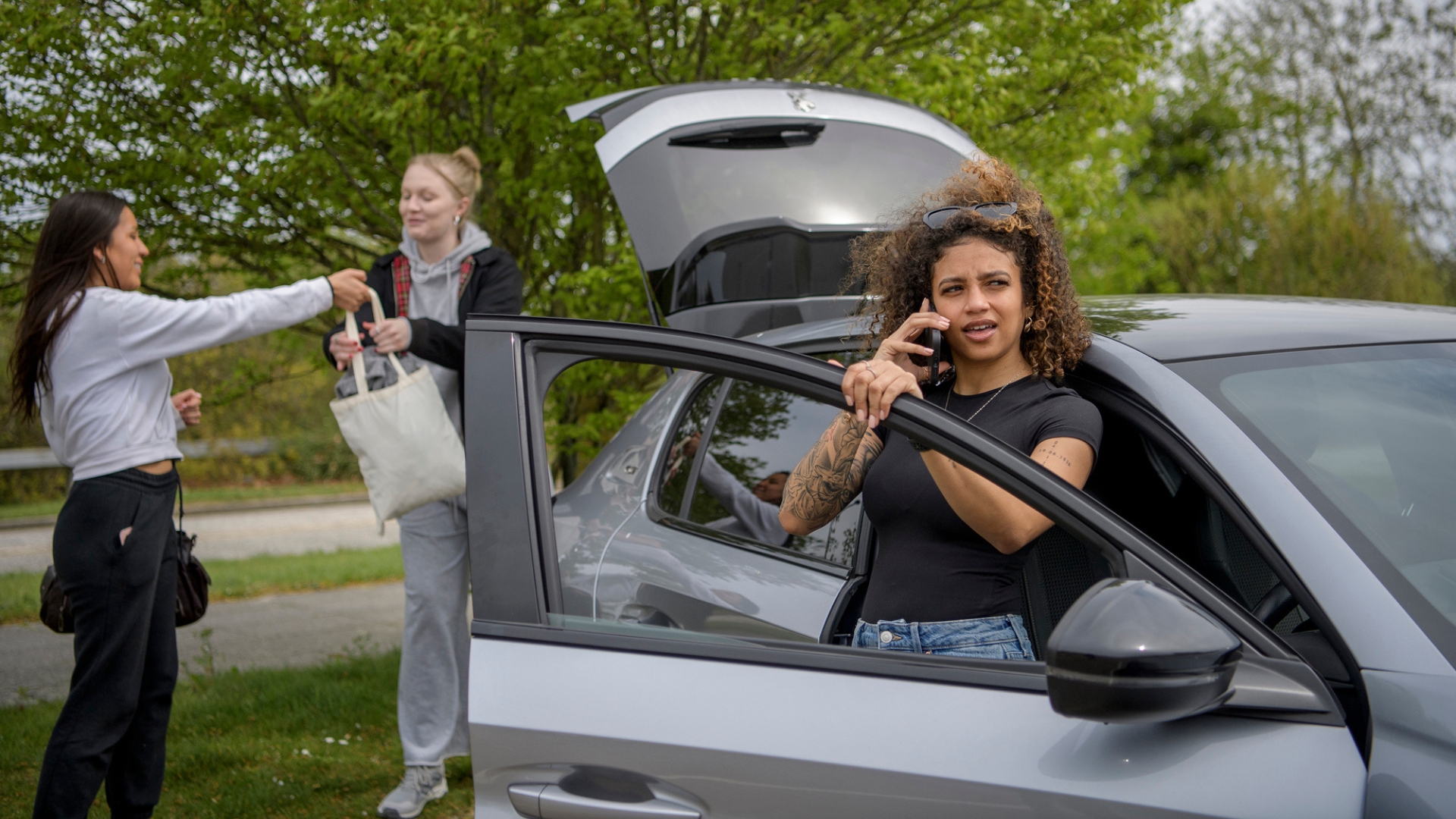 Person standing next to a car with the door open talking on a phone, with two people behind the car putting a bag into the open boot.