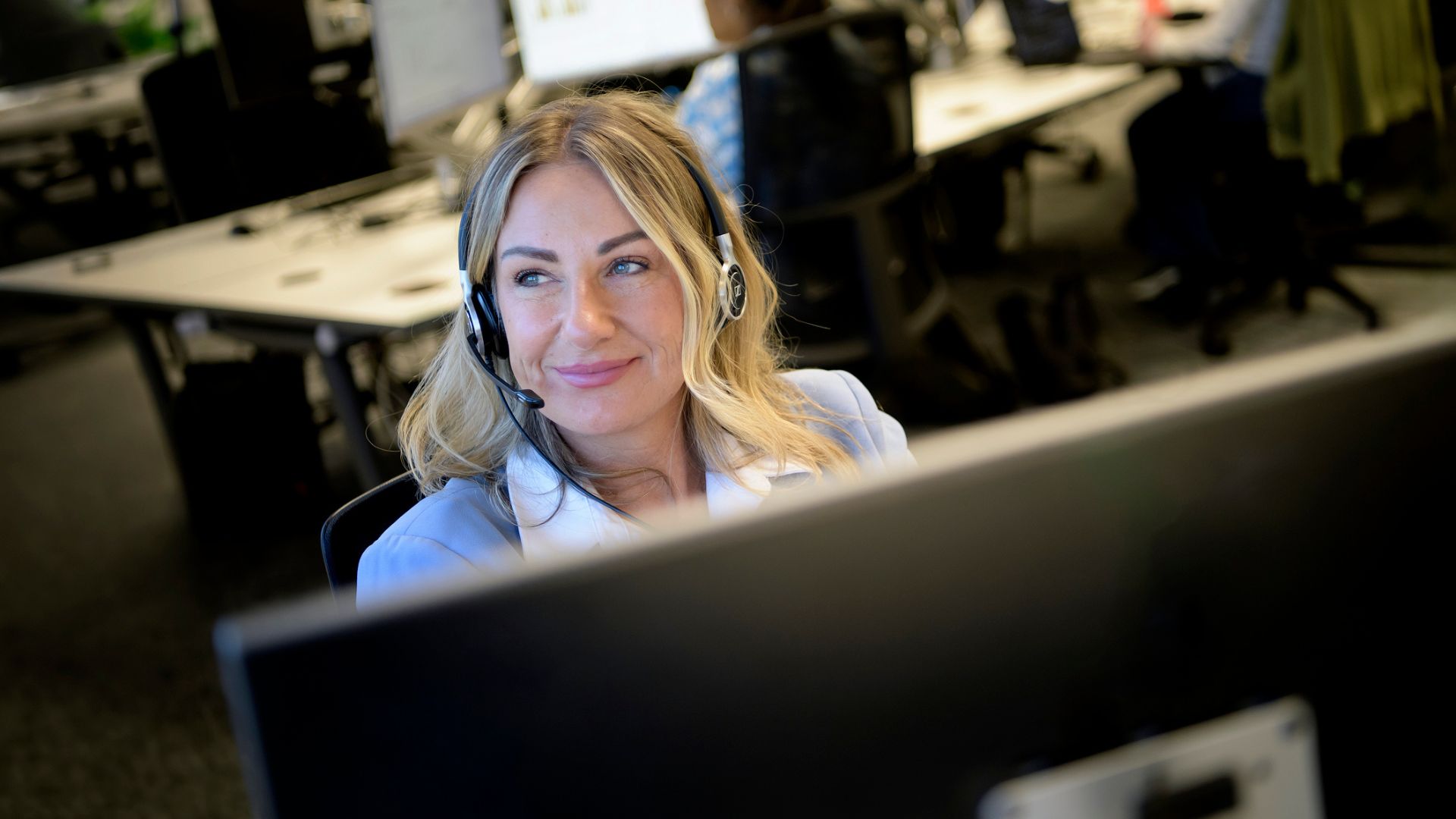 Blond woman pictured wearing call centre headset, sitting behind computer.