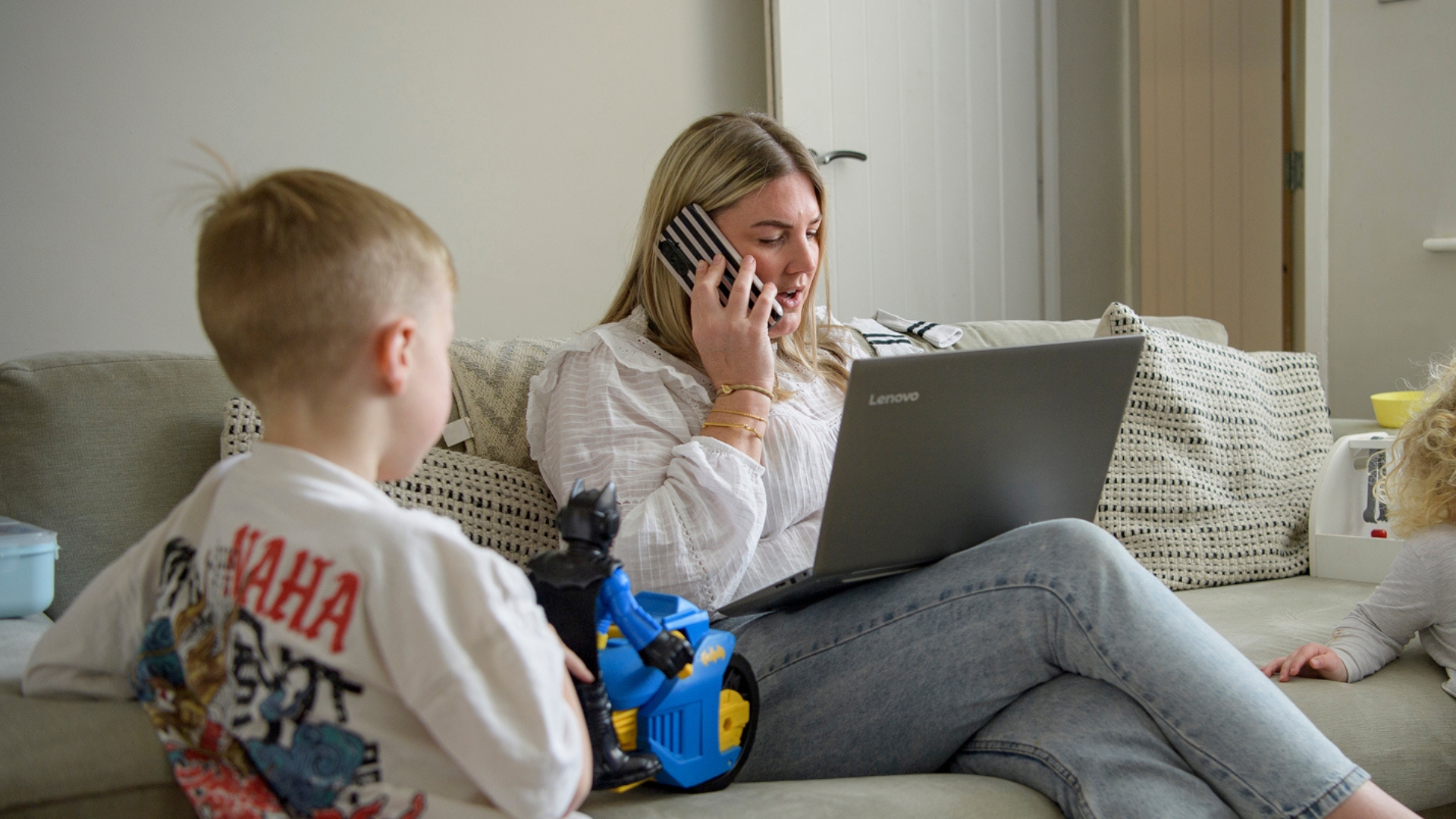A person sitting on a sofa using a laptop while talking on a phone, with a child nearby.
