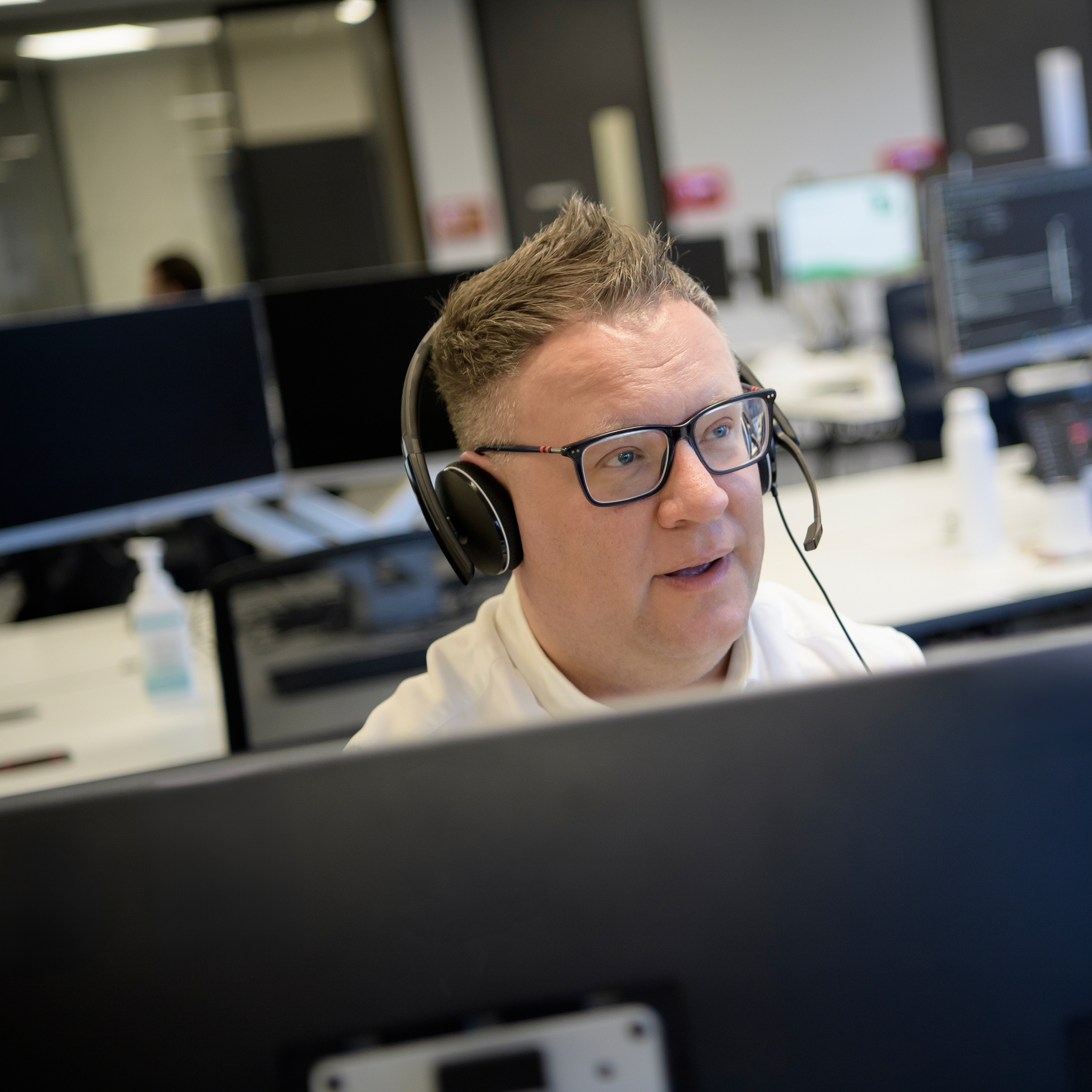 A person wearing a headset working at a computer, with other desks and screens in the background.