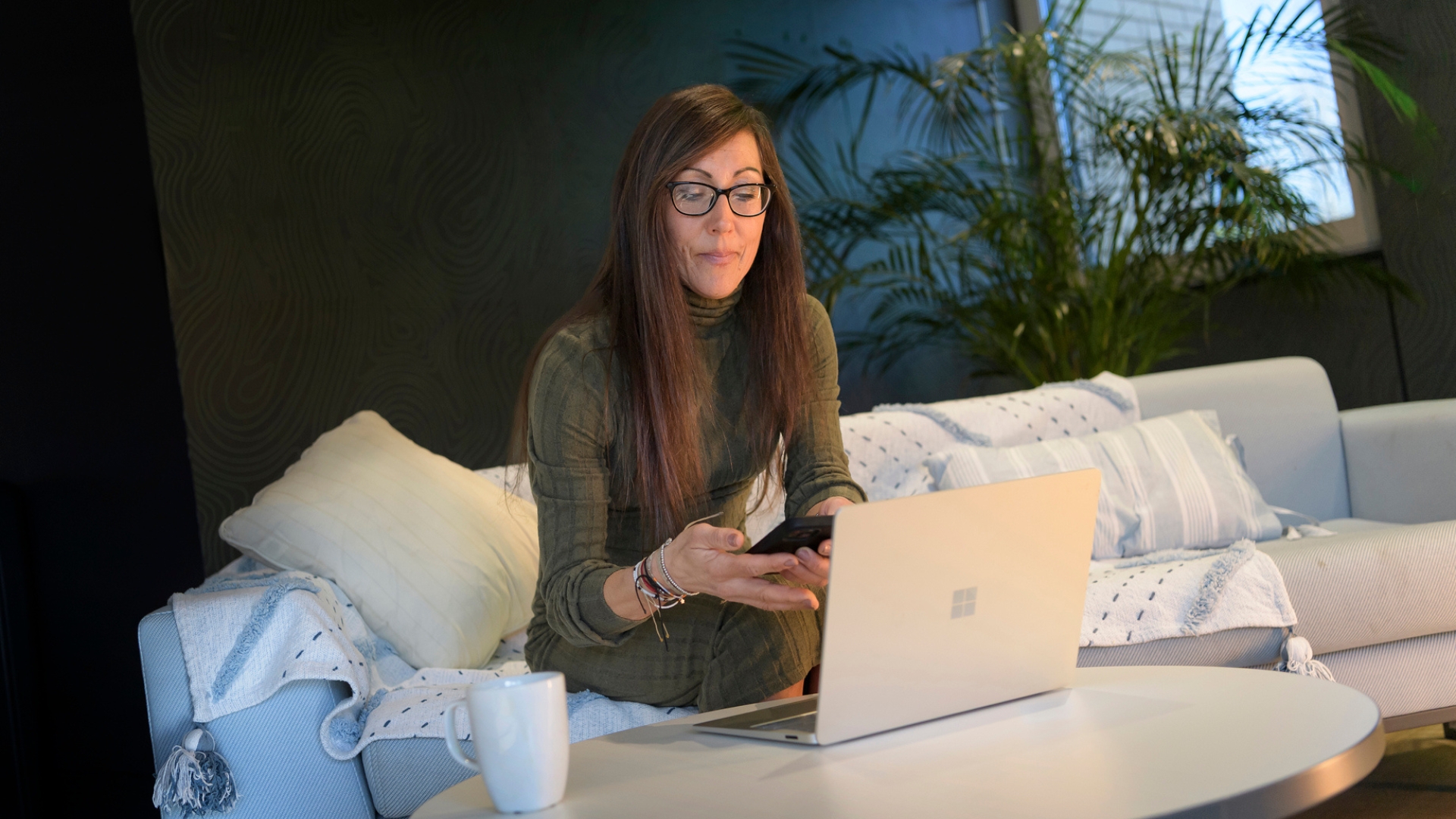 A person on a sofa using a laptop while holding a mobile phone, managing a claim online.