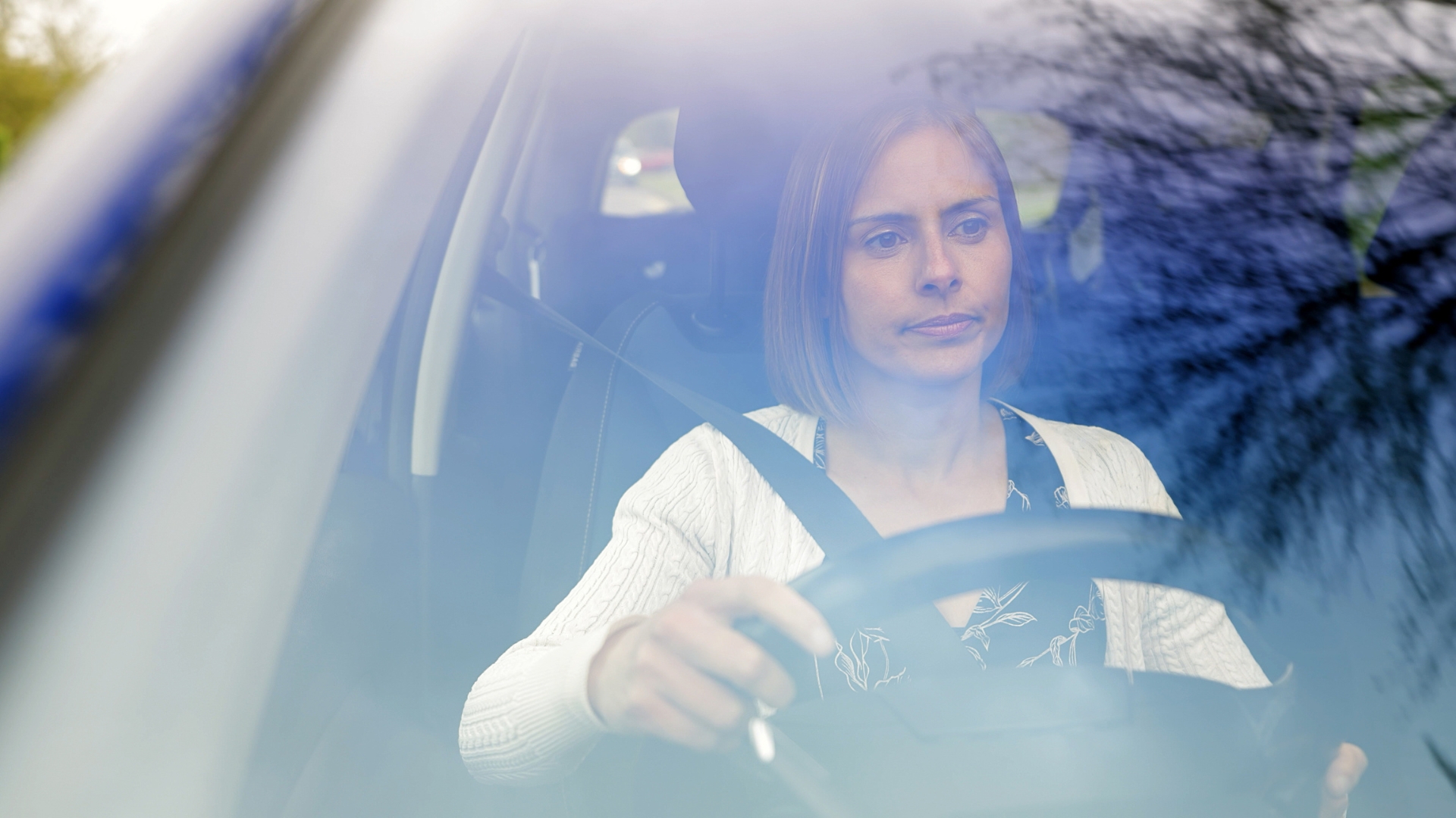 Person driving a car, seen through the windscreen, wearing a seatbelt.