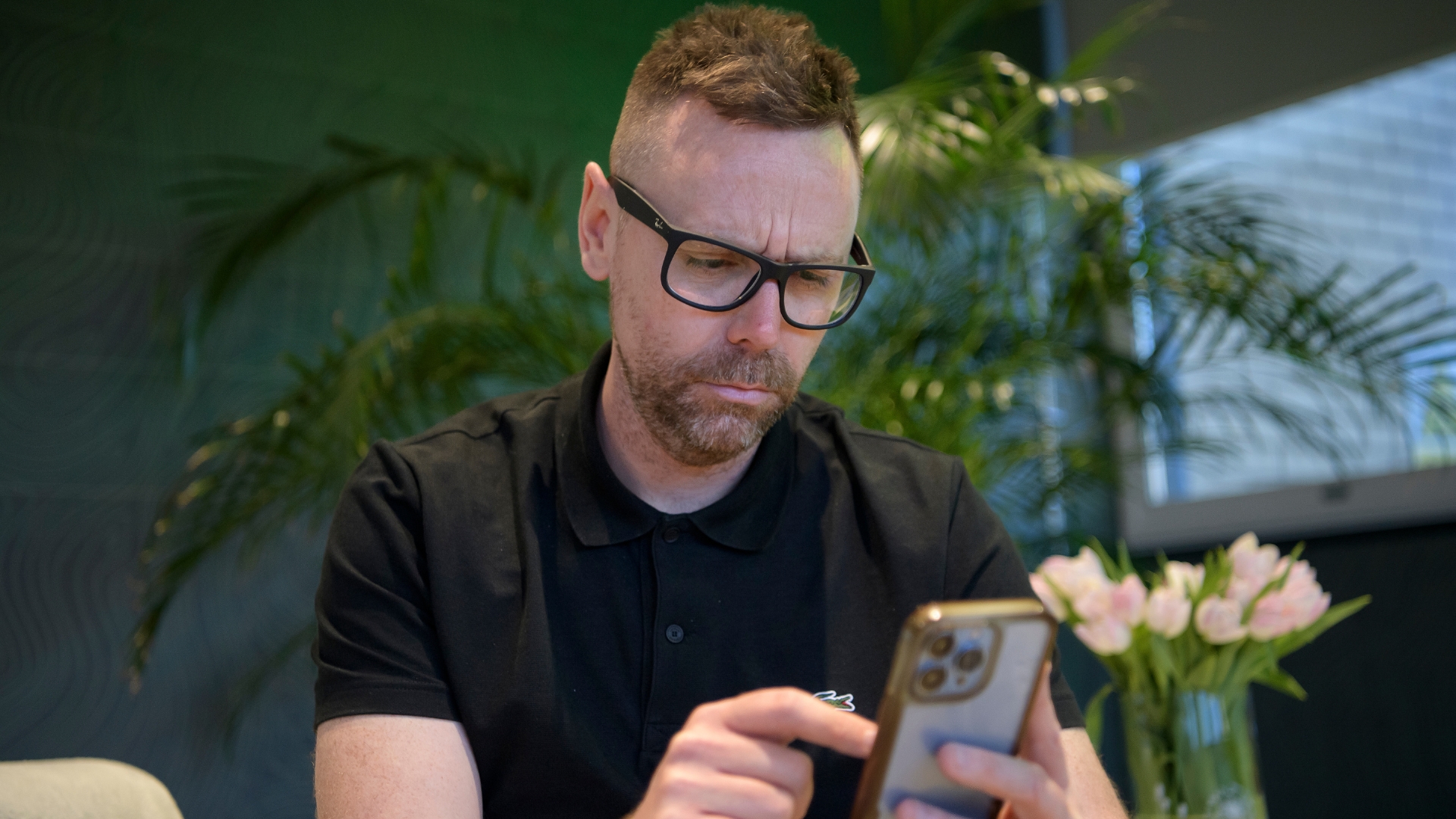 A person using a smartphone while sitting at a table indoors, looking confused.