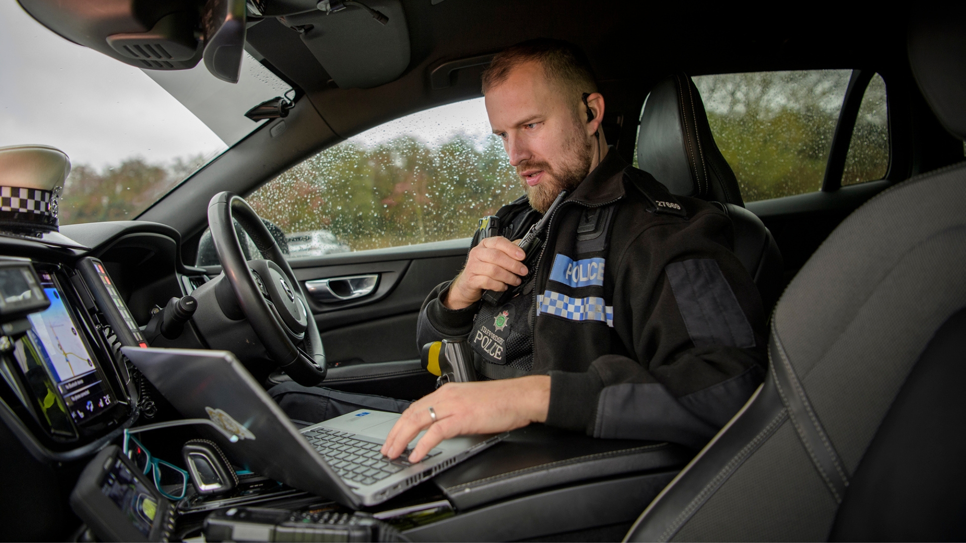 Police officer in a roads policing vehicle using a laptop while speaking into a radio.