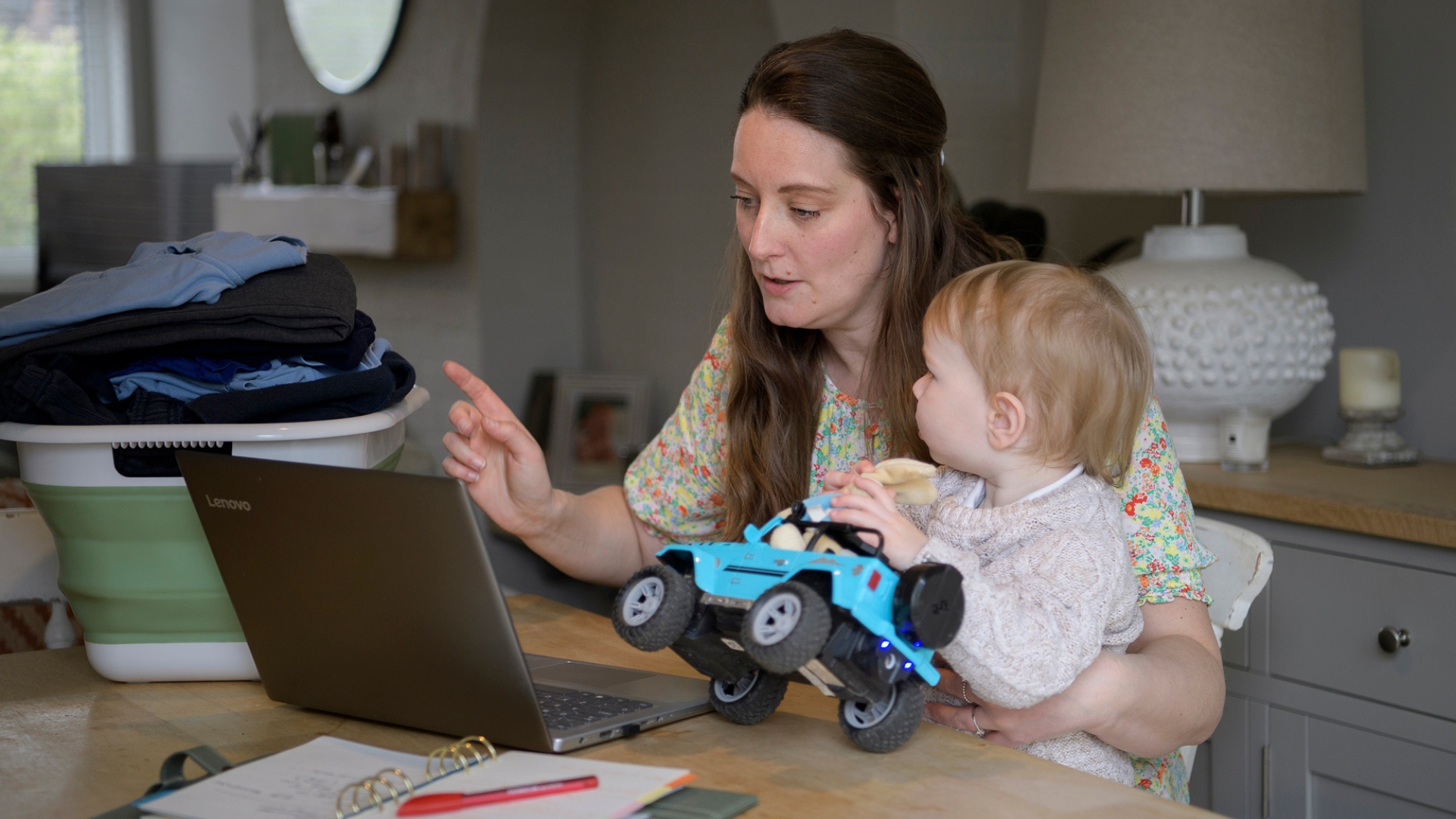 An adult using a laptop at a table with a child on their lap, with laundry beside them.