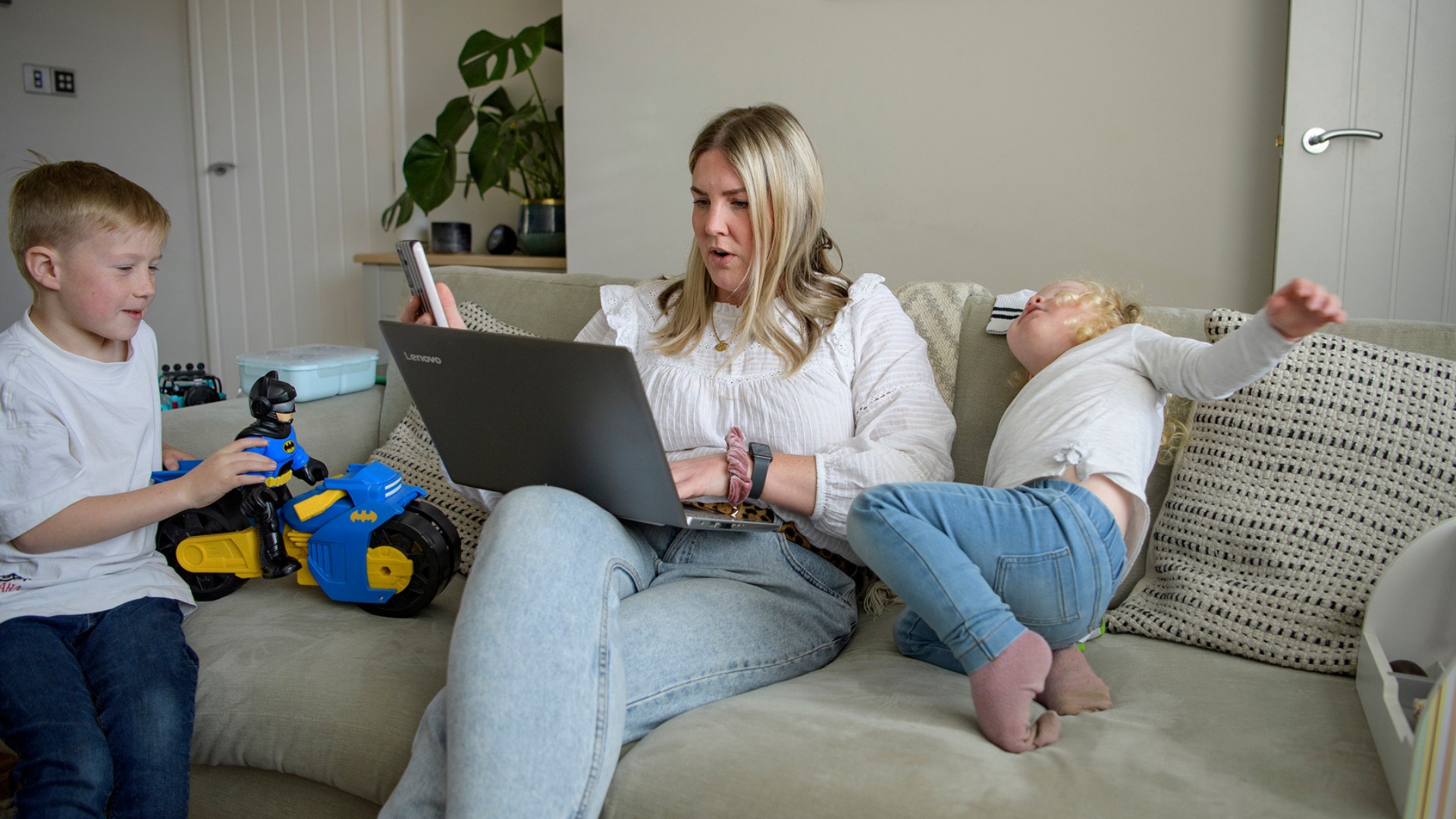 An adult using a laptop and mobile phone on a sofa in a living room, with two children nearby.